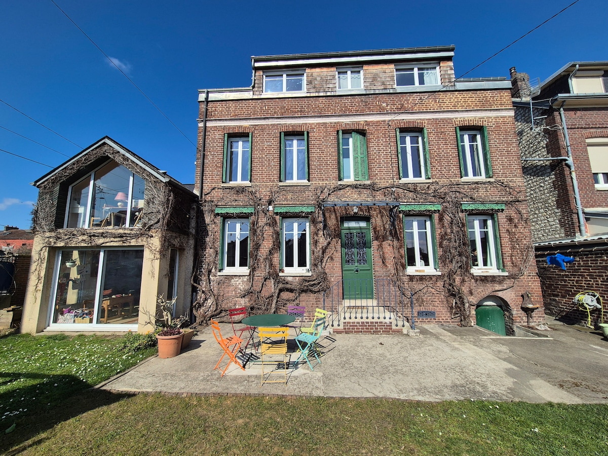 The exterior of a charming town house is visible, featuring a facade adorned with climbing plants. Brightly colored chairs surround a circular table on a grassy area, offering ample outdoor seating. Large windows allow for natural light, and the sky is clear and blue.