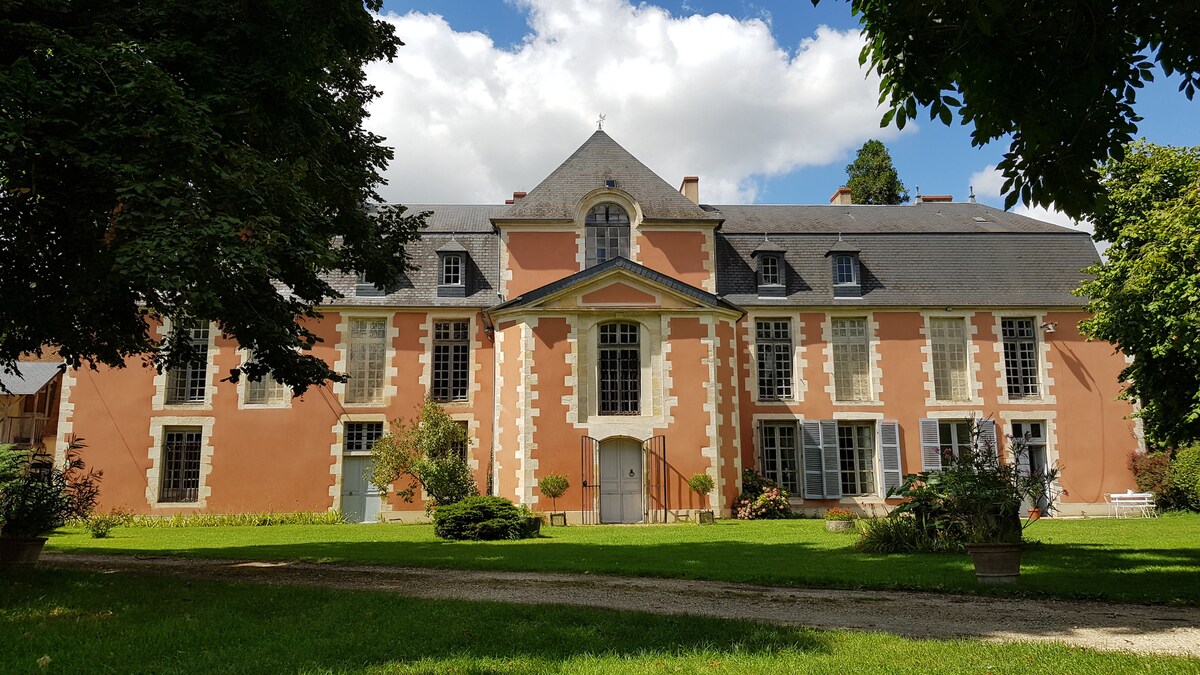 The castle's exterior features a blend of pale pink and white stone, complemented by large windows. Lush green landscaping surrounds the building, providing a serene atmosphere. The structure showcases architectural elements from the 17th century, emphasized by a central entrance and symmetrical design.