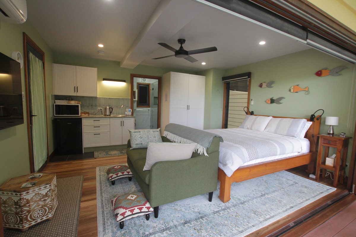 A bright guesthouse interior features a spacious bed, complemented by soft linens. A kitchenette is equipped with essential appliances, while a comfortable seating area offers a cozy spot to relax. Light wood flooring and calm green walls enhance the overall serene atmosphere.