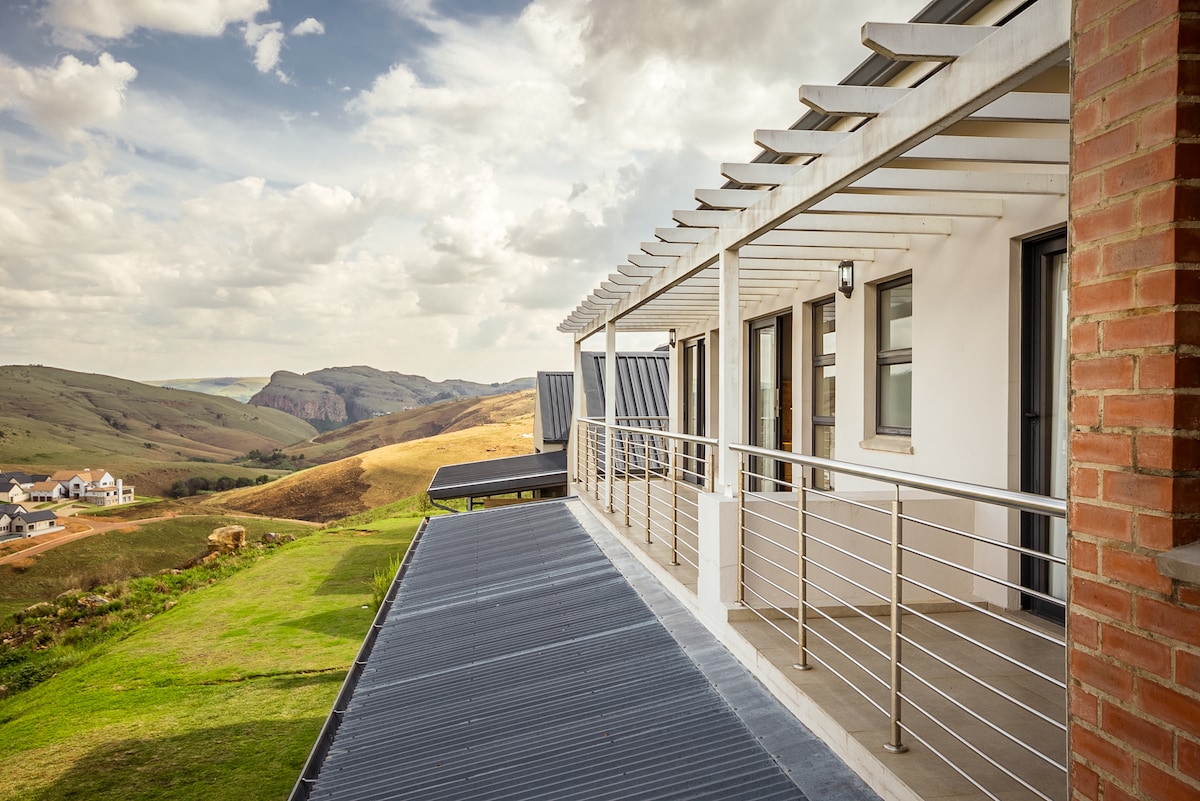 A spacious balcony is shown, featuring a modern metal railing and multiple sliding doors. The landscape reveals rolling hills and distant mountains under a partly cloudy sky, providing a scenic backdrop. The balcony's clean lines offer a contemporary aesthetic.