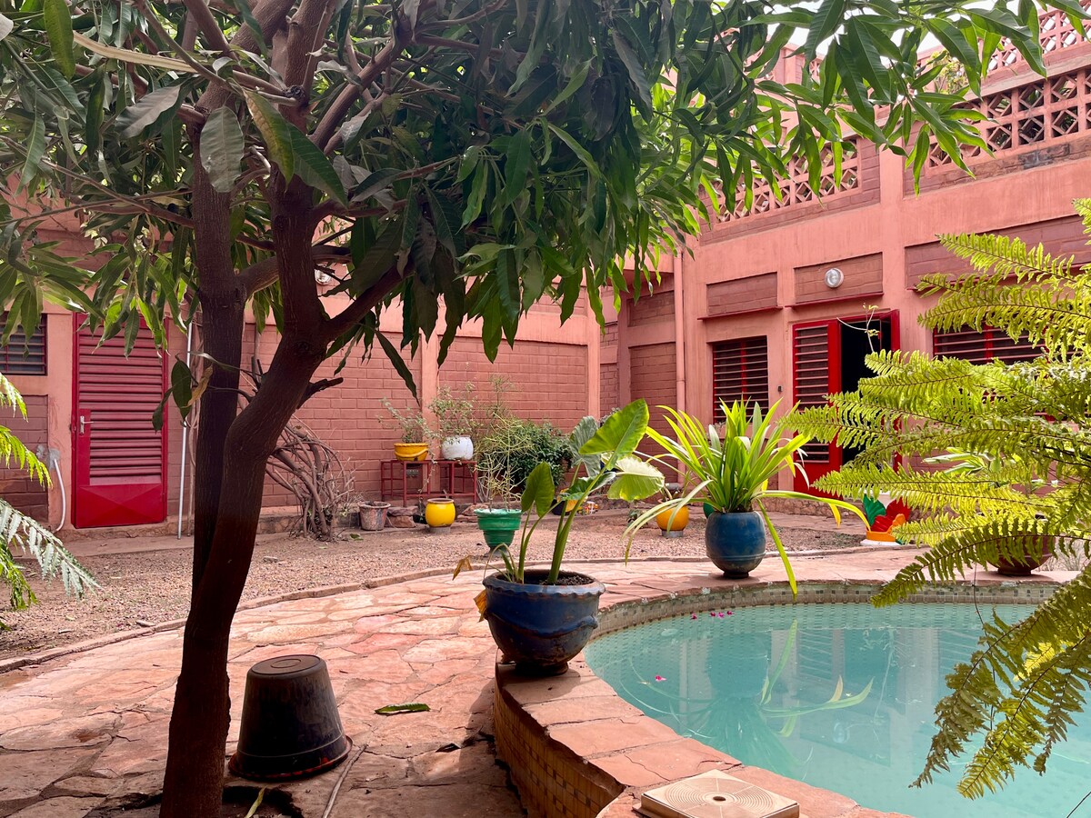 The image captures a tranquil courtyard featuring a small pool surrounded by vibrant greenery. A tree casts shade over the area, while potted plants add bursts of color. The rustic red walls of the duplex complement the serene outdoor space.