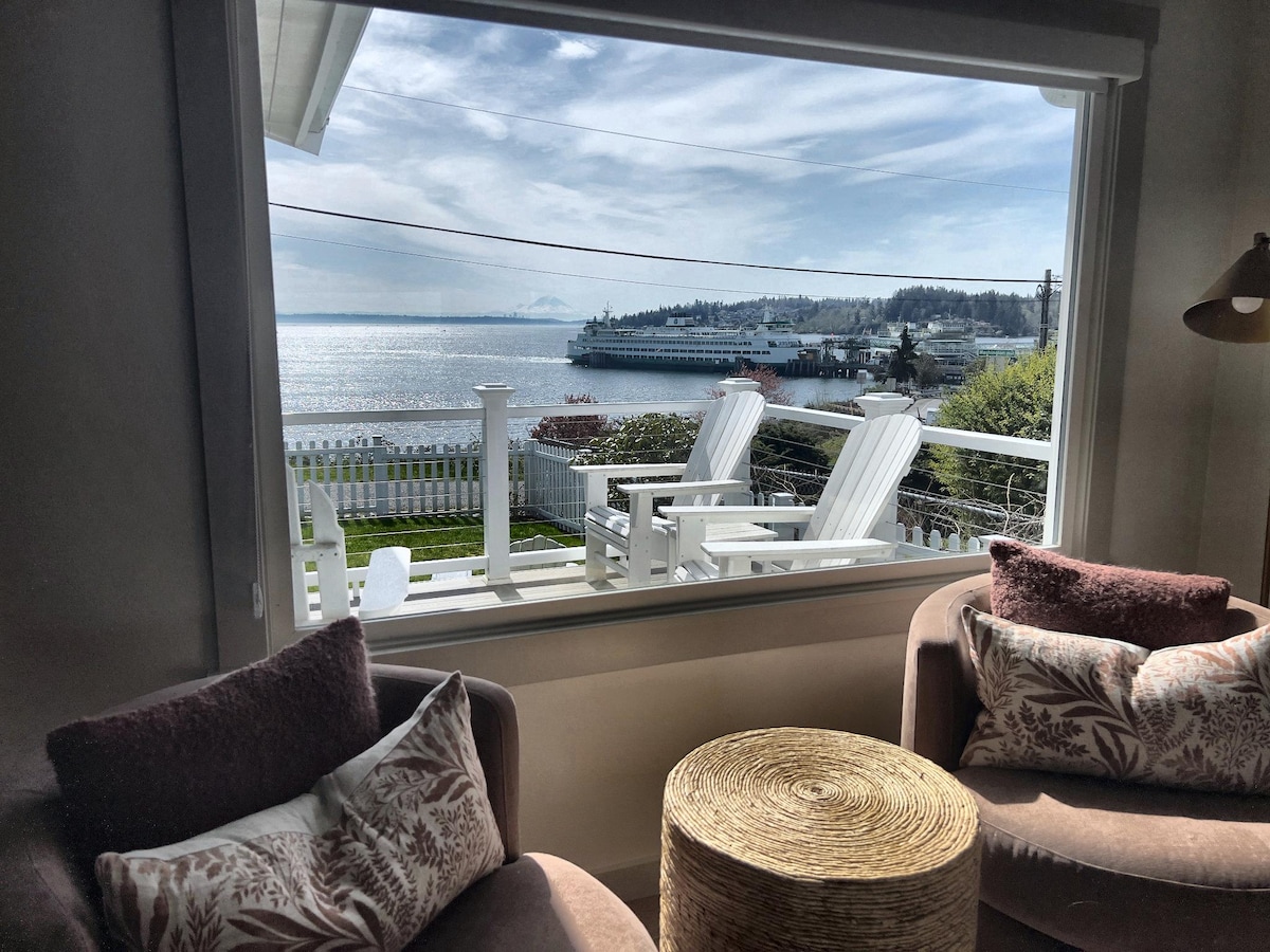 The living room offers a large picture window framing a full view of the water. Two lounge chairs are positioned beside a woven coffee table, creating a cozy space for relaxation. Sunlight fills the room, highlighting the serene outdoor scenery.