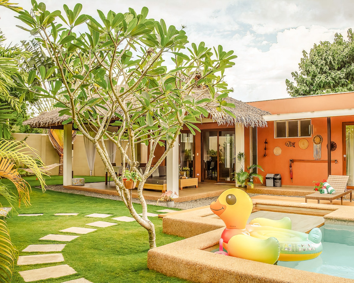 A vibrant outdoor scene features a private pool surrounded by lush greenery and a tropical garden. A playful yellow inflatable floats in the pool, while a thatched-roof terrace offers a shaded area. Stone pathways lead through the manicured lawn, creating inviting spaces for relaxation.