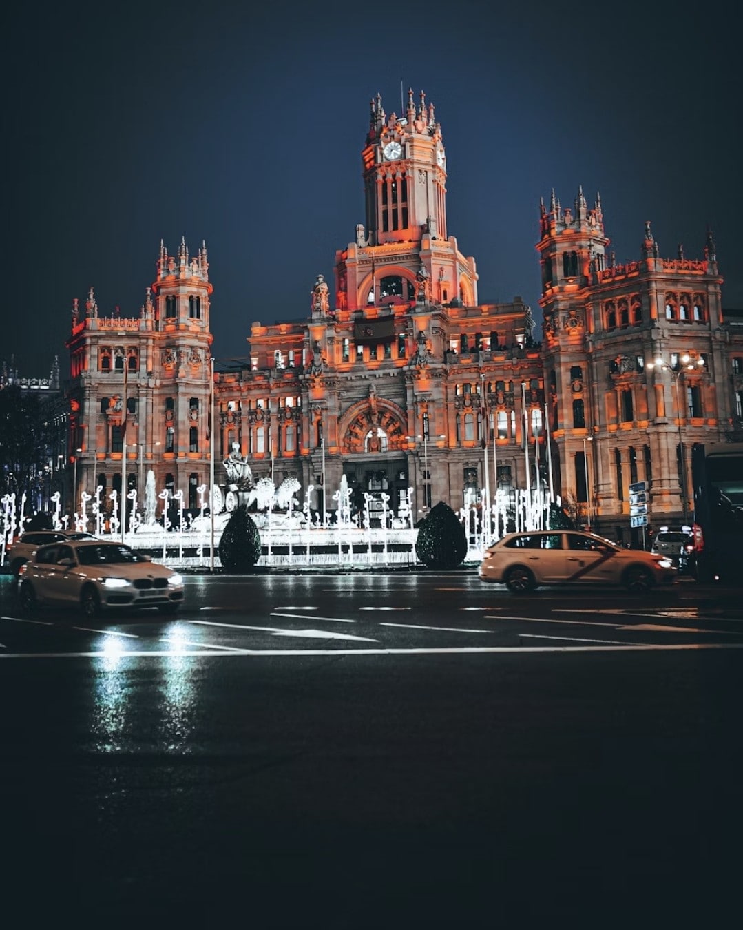 An illuminated historic building stands majestically against a night sky. Its elaborate architecture features towers and detailed carvings, while water fountains dance in front, reflecting the building's warm lights. Cars are seen passing by, creating a bustling city atmosphere.