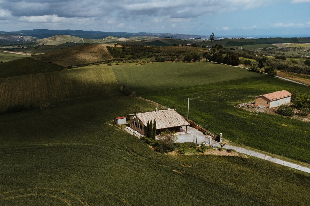 An aerial view showcases a rustic villa surrounded by expansive green fields and rolling hills. The house features a gentle sloping roof and is positioned along a narrow gravel road. Nearby, a smaller structure is visible against the landscape, highlighting the serene rural setting.