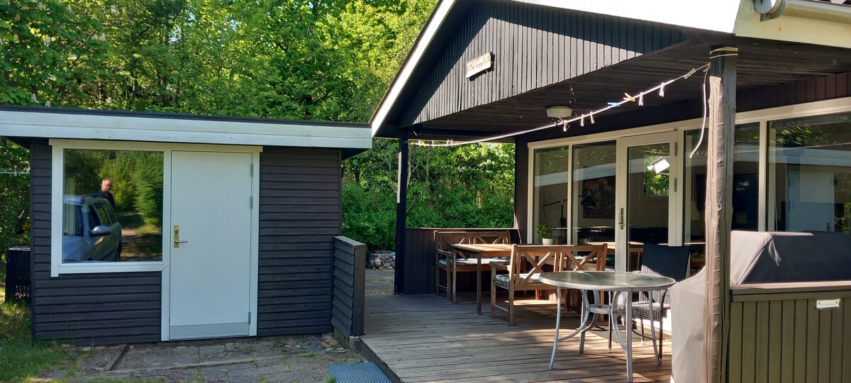 A wooden cabin is positioned alongside a small annex, both surrounded by lush greenery. The main structure features large glass windows and a spacious terrace furnished with a dining table and chairs. A barbecue area is visible, enhancing outdoor leisure options.