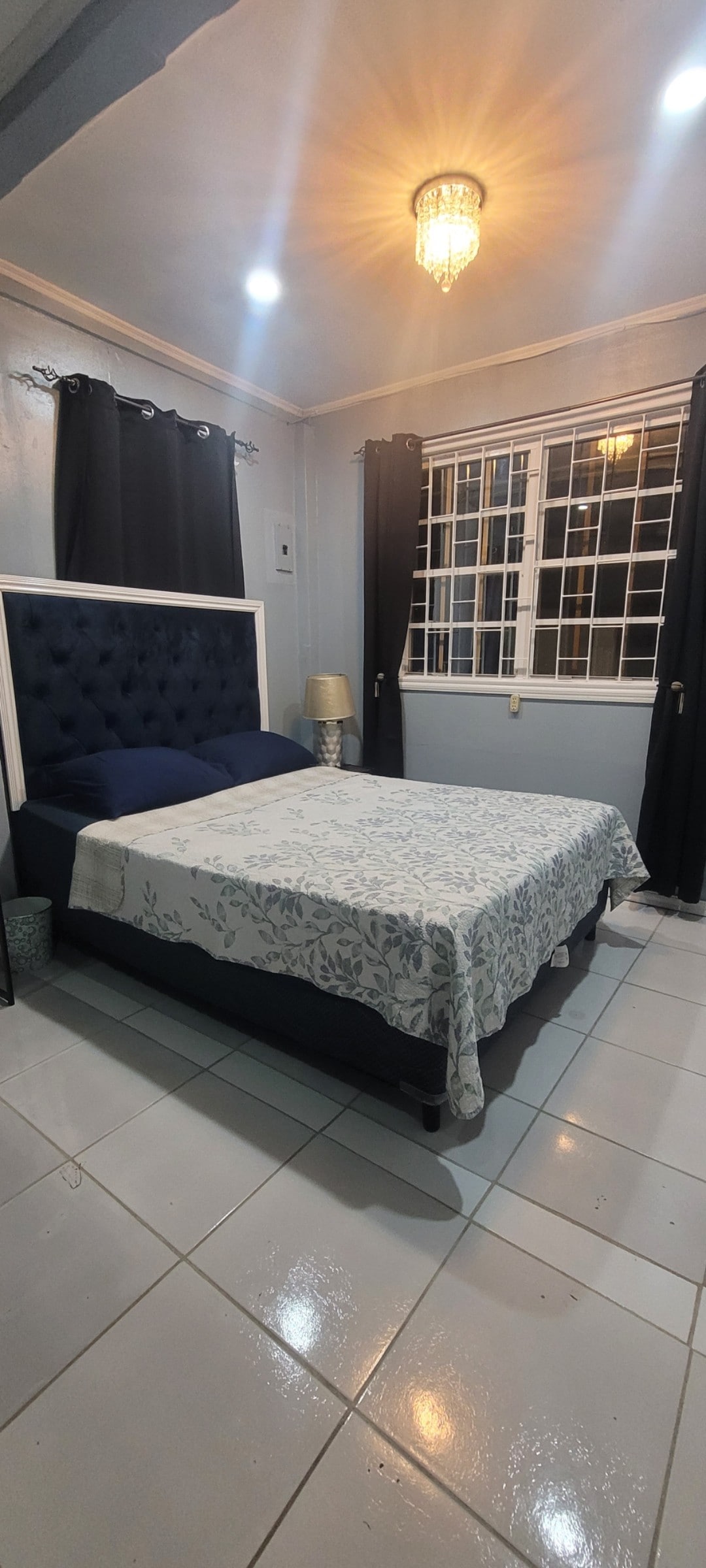 A spacious sleeping area features a plush bed with a decorative light blanket. Soft lighting from a chandelier and wall lamps illuminates the room, while large windows are framed by dark curtains, creating a cozy ambiance.