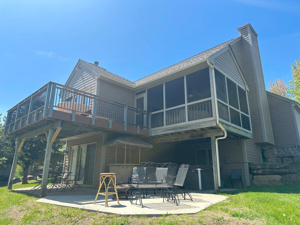 The exterior of a spacious two-story house is shown, featuring a large screened porch and a deck above. A patio area with multiple chairs is visible, surrounded by a green lawn. Sunlight casts a bright glow on the structure.