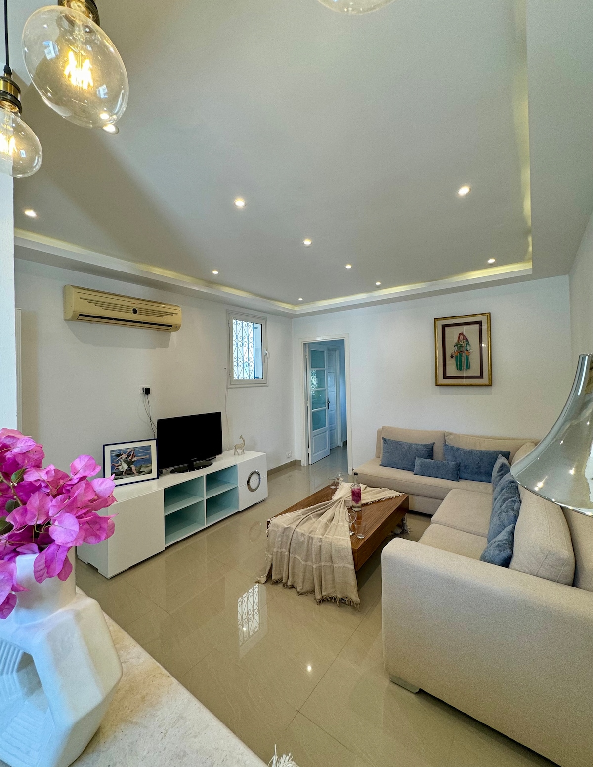 A well-lit living room features a light-colored sectional sofa paired with a wooden coffee table. A television is positioned on a modern white cabinet. Decorative flowers add a touch of color. Soft lighting fixtures are integrated into the ceiling, enhancing the inviting ambience.