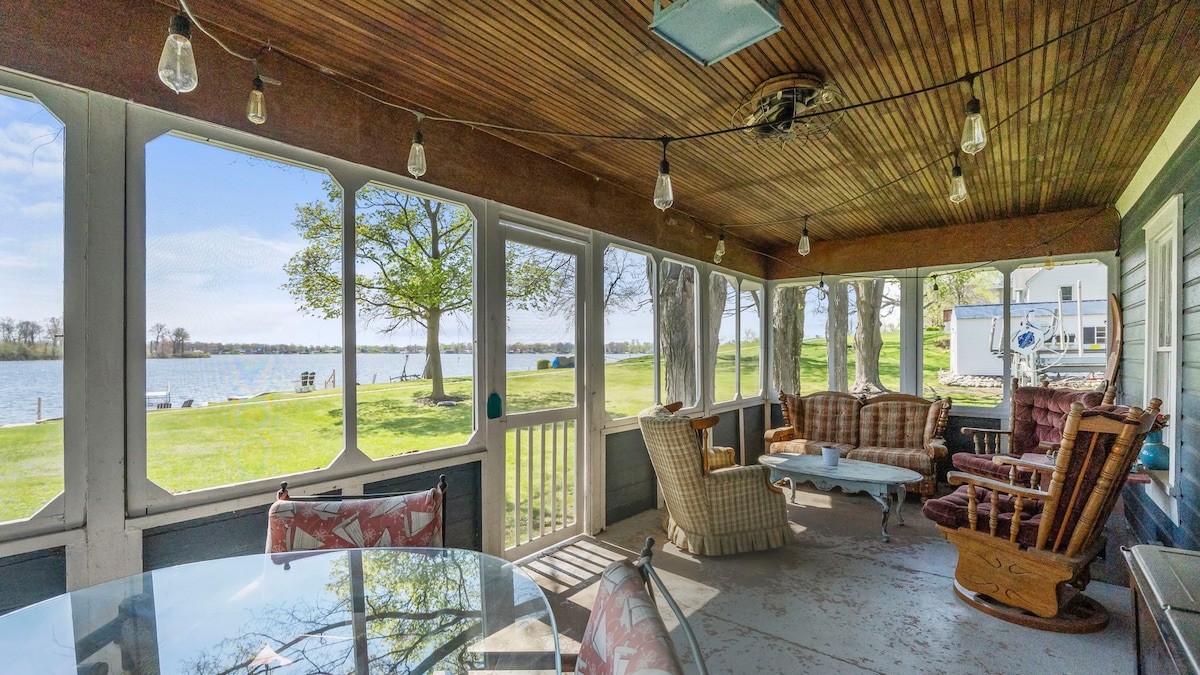 The screened-in porch offers a comfortable seating area with a wooden rocking chair and a cozy couch, framed by large windows providing a view of the lake. A small table sits in the center, and lights hang overhead, creating a pleasant ambiance.