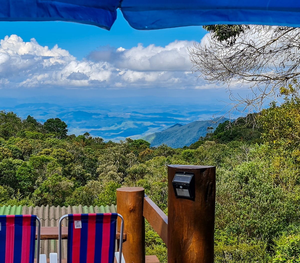 A spacious deck features comfortable chairs complemented by vibrant striped fabric. The view extends across lush greenery and distant mountain peaks under a clear blue sky, creating a serene atmosphere for relaxation. A mailbox is positioned nearby, blending with the natural surroundings.