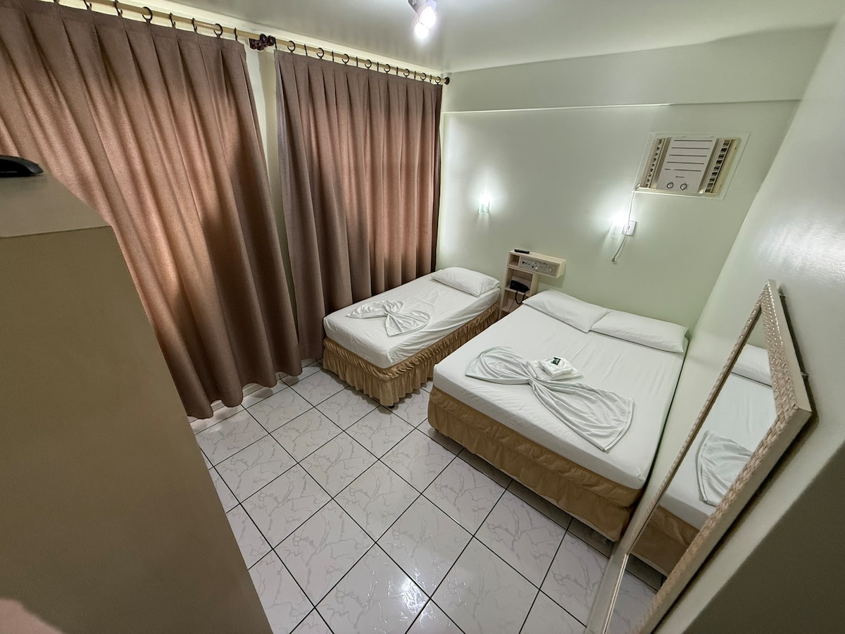 The sleeping area features two spacious beds with neatly arranged linens. A full-length mirror reflects the room. Natural light enters through curtains, adding warmth, while air conditioning and a minibar are available for comfort.