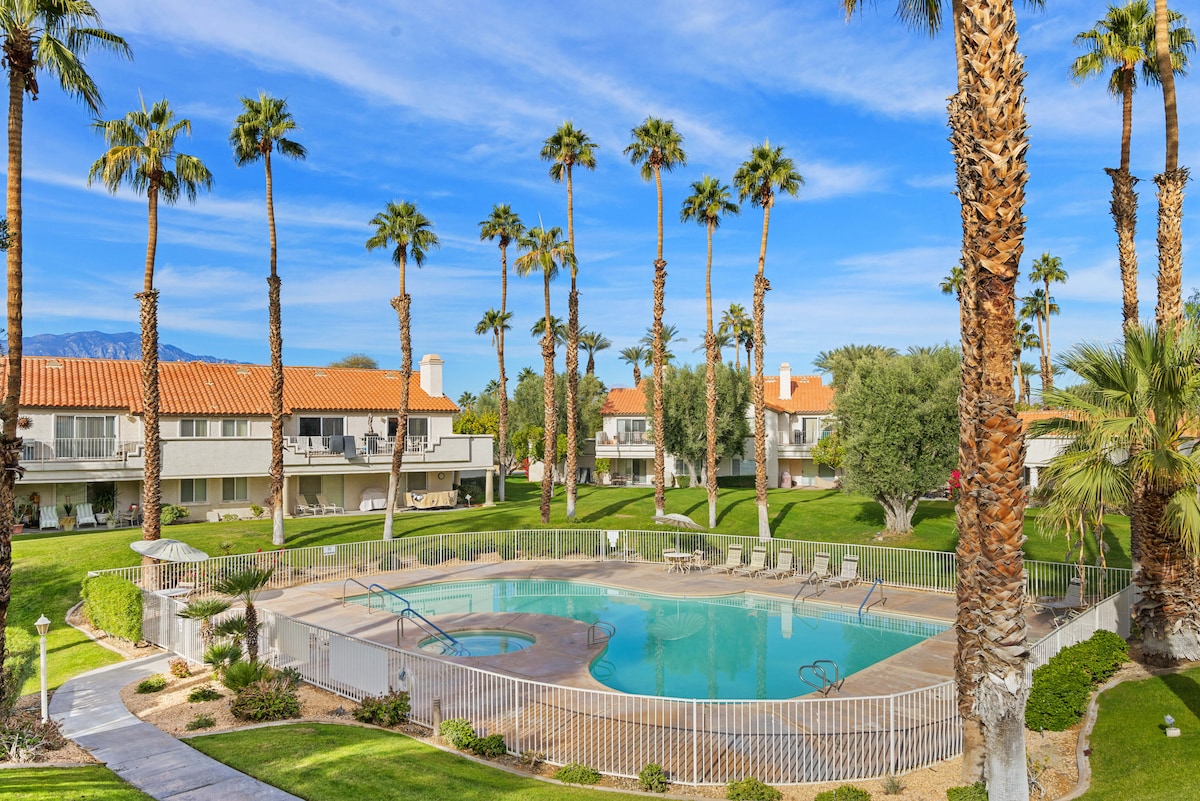 A tranquil swimming pool is surrounded by vibrant palm trees and well-maintained green landscaping. The pool area features lounge chairs arranged neatly around the water. In the background, the villa's welcoming architecture is visible under a clear blue sky, enhancing the outdoor space.