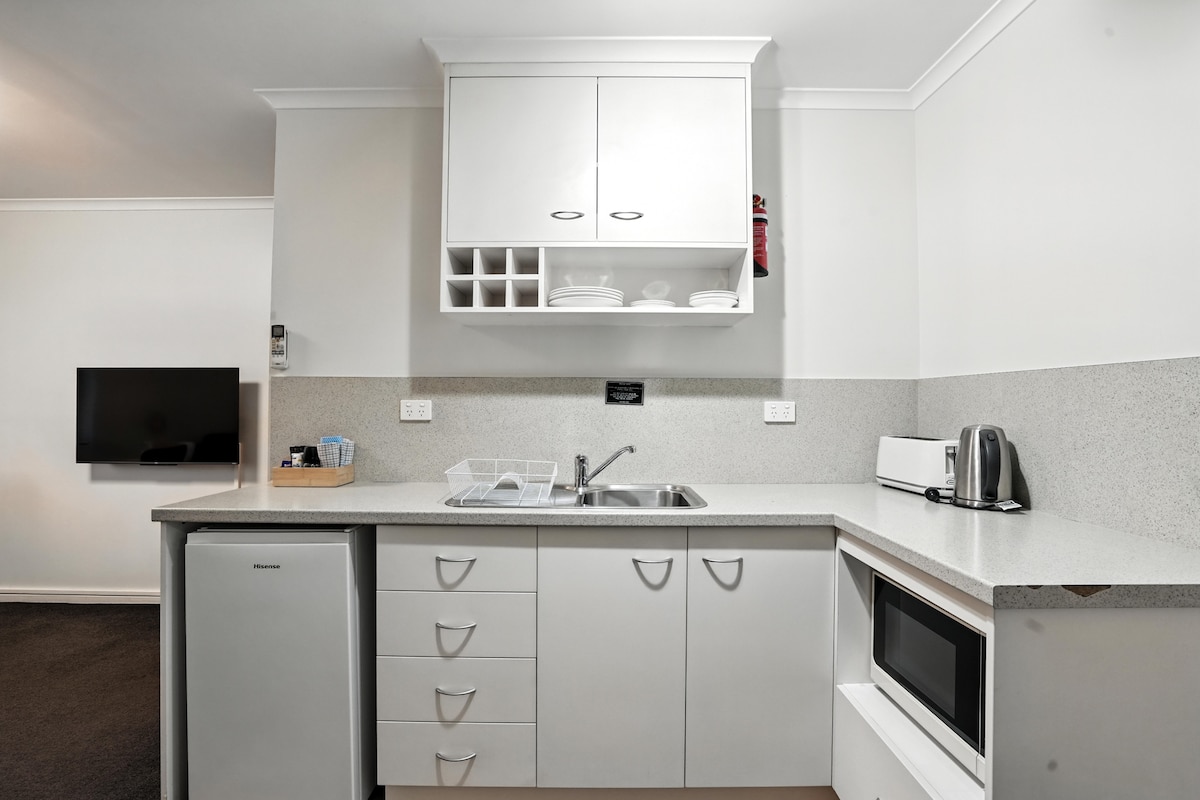 A compact kitchen area is presented, featuring essential appliances including a mini fridge, microwave, kettle, and toaster. White cabinetry provides ample storage, while dishware is neatly arranged on open shelving. A sink is positioned near the countertop, with a flat-screen television mounted on the adjacent wall.