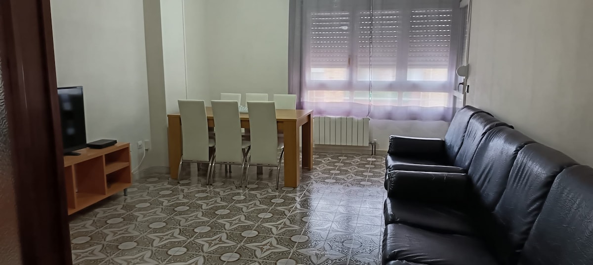 A bright living area is shown, featuring a black leather sofa and a wooden dining table with white chairs. Natural light filters through sheer curtains, illuminating the patterned tile flooring. A television is positioned on a small wooden stand against the wall.