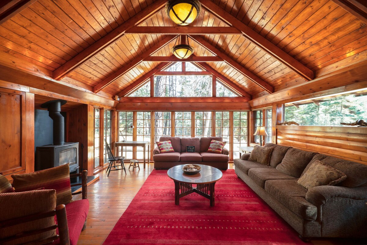 A spacious living area features a wooden ceiling with exposed beams, large windows offering views of the surrounding nature, and a cozy seating arrangement including a sofa and armchair. A round coffee table sits in the center, complemented by a decorative rug underneath.