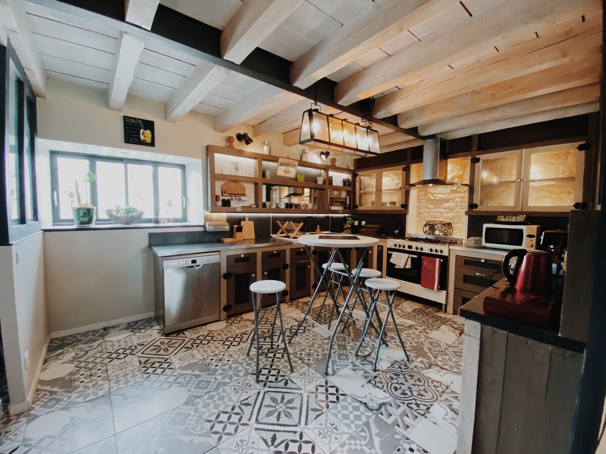 The kitchen features exposed wooden beams and a mix of modern and rustic details. Stainless steel appliances are complemented by wood accents. A central island is surrounded by three stools, and large windows provide natural light, highlighting the patterned floor tiles.
