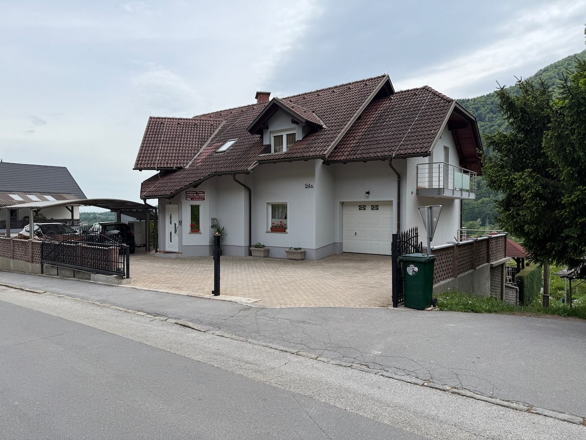 A two-story guest house is shown, featuring a red-tiled roof and a white exterior. The building is set on a paved area with pots of flowers at the entrance. A carport is visible on the left, alongside green trees and a hillside in the background.