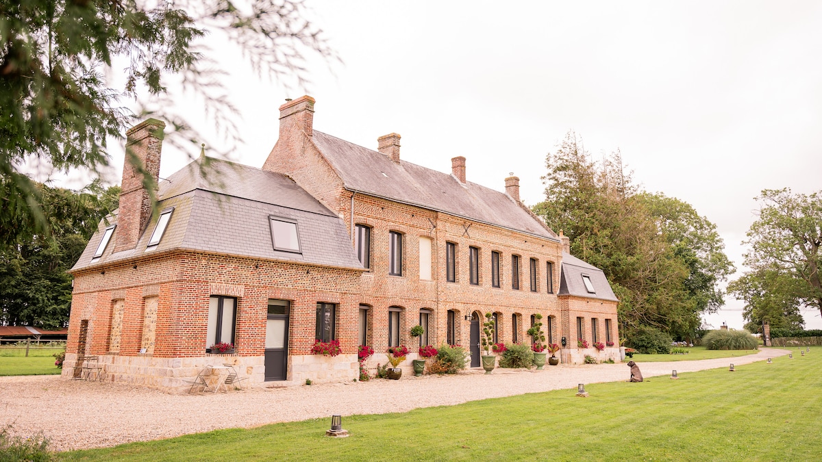 A historic brick manor, characterized by its distinct architecture and large windows, stands in a manicured lawn. A gravel pathway leads to the entrance, bordered by greenery and flower arrangements. The setting is surrounded by mature trees, creating a serene atmosphere.