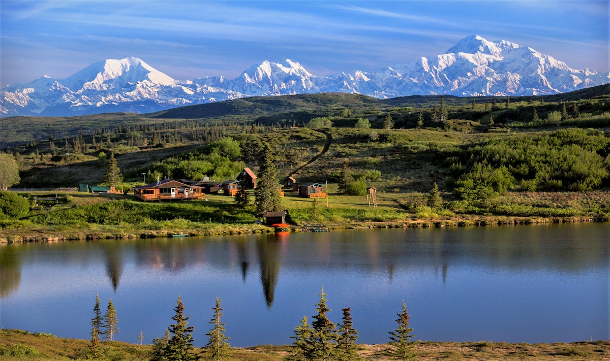 The serene lodge is situated near a lake, surrounded by lush greenery and majestic snow-capped mountains in the background. Reflections of the landscape are visible on the calm water, creating a harmonious blend of nature and tranquility.