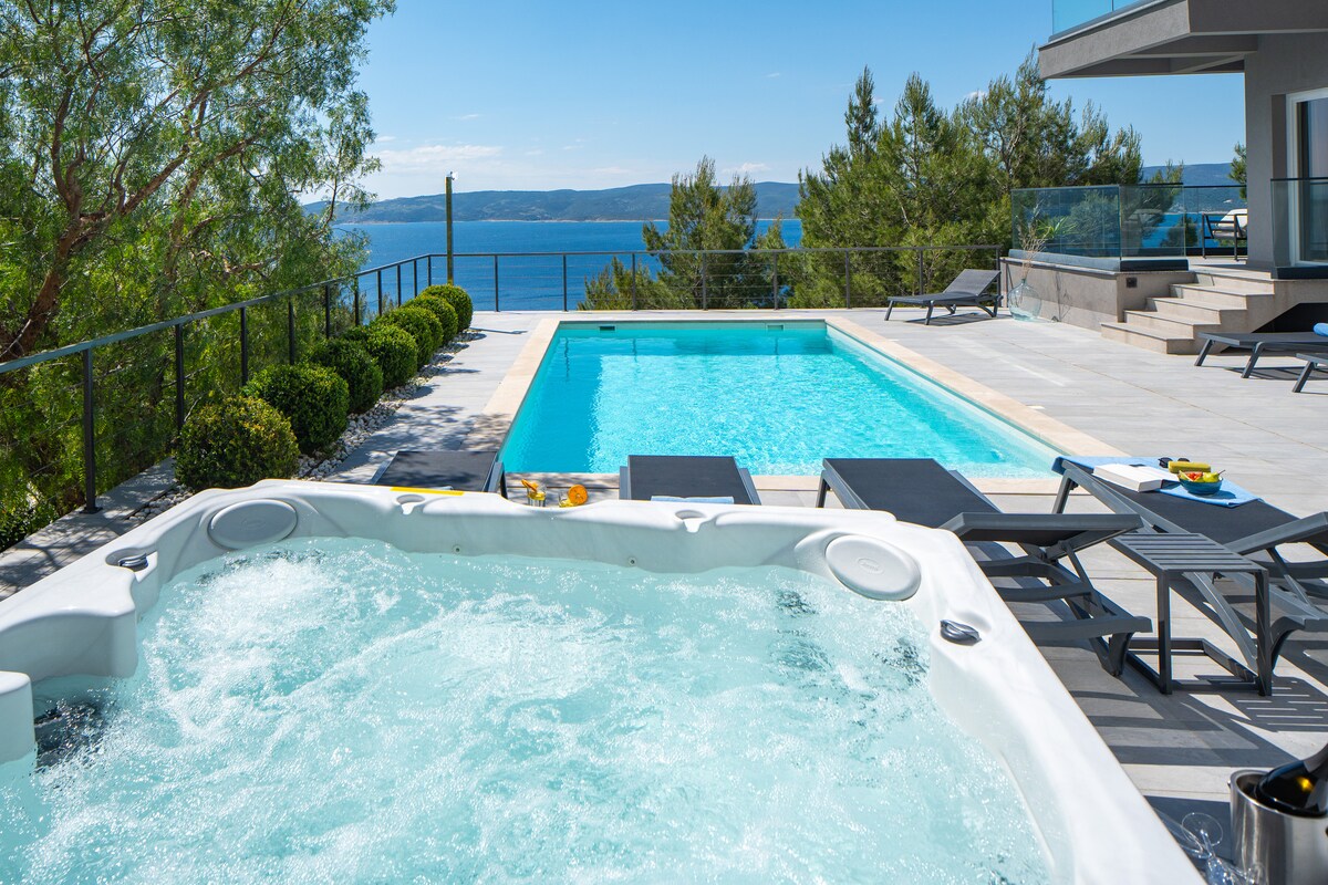 An outdoor area features a private pool alongside a whirlpool, both with sweeping views of the sea and mountains. Loungers positioned by the pool invite relaxation under the sun, while neatly trimmed hedges provide a charming border to the space.