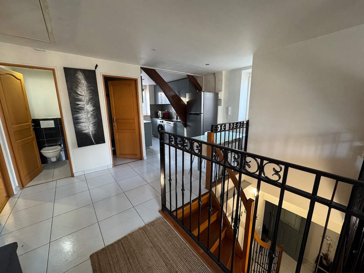 A spacious entryway features a wooden staircase leading to an upper level. Natural light fills the area through large windows. The design includes tiled flooring and wooden doors that open to various rooms, while an airy rug enhances the floor space.