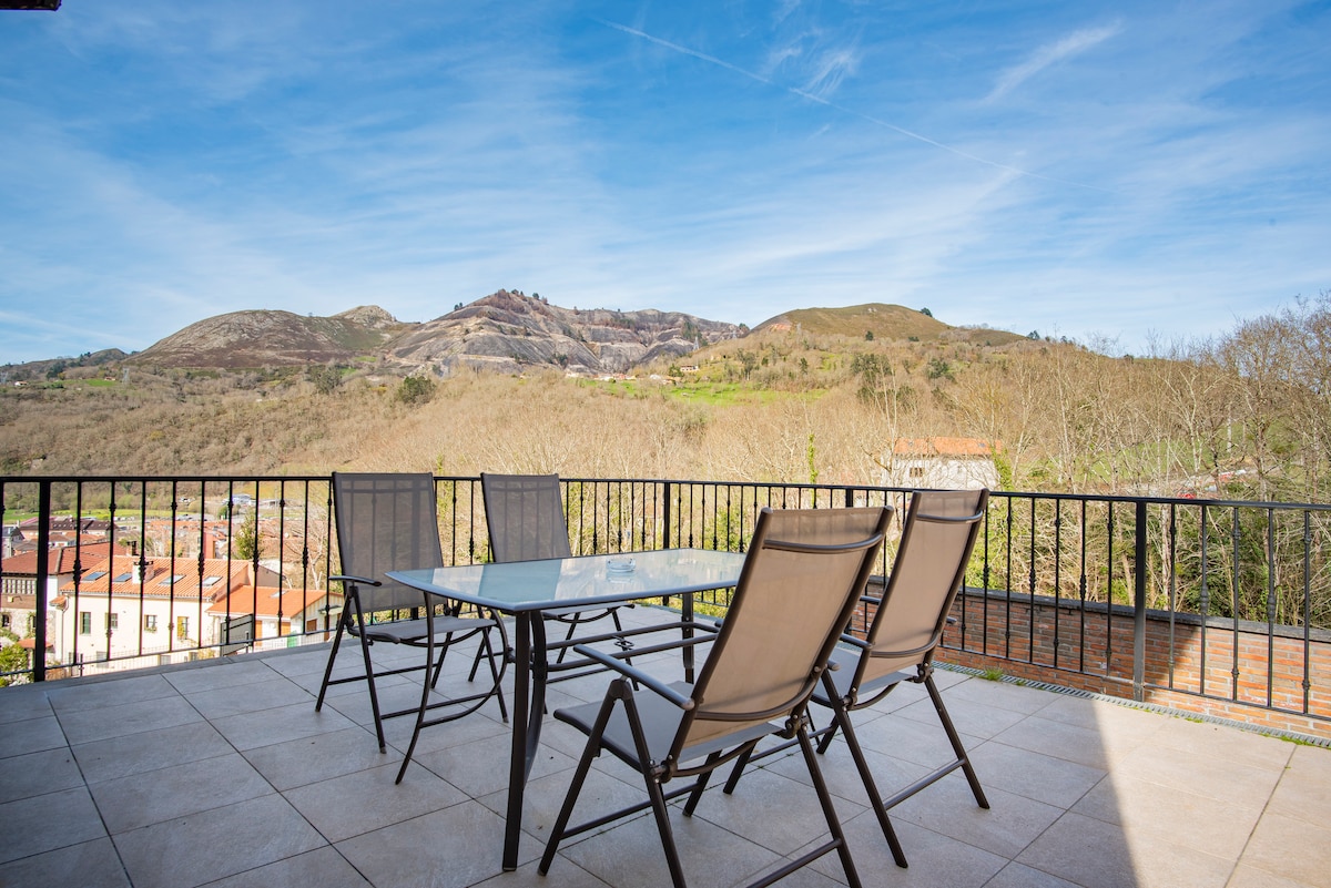 An outdoor terrace features a glass-top dining table surrounded by several chairs. A panoramic view of surrounding hills and mountains is visible in the background, with the tranquil landscape creating a serene setting. The terrace is designed for outdoor dining and relaxation.