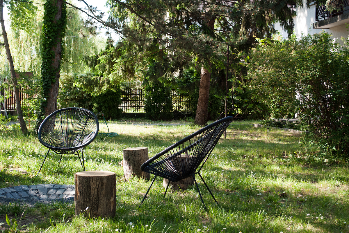 A tranquil outdoor space features two black, woven chairs positioned on a grassy area. Nearby, two wooden stumps serve as natural tables. Lush greenery surrounds the scene, providing a peaceful atmosphere suitable for relaxation.