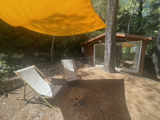 Two sun loungers are positioned underneath an orange shade sail, surrounded by a natural landscape. The chalet, crafted from wood, is set in the background, with large windows allowing for ample light. Pine trees provide a serene backdrop to this outdoor space.