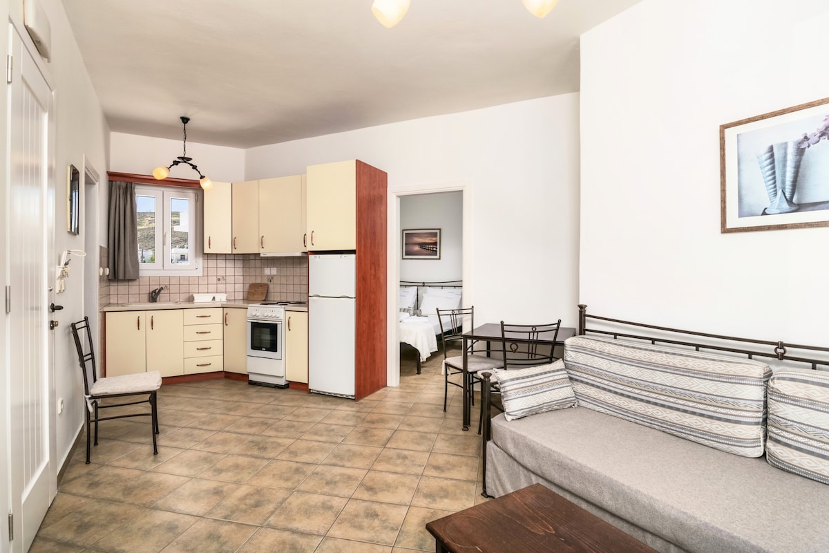 A light-filled living space features a comfortable sofa against a white wall, complemented by a wooden coffee table. The kitchen area is visible with modern appliances and a dining table set for four. Neutral tiles enhance the open layout, offering a welcoming atmosphere.