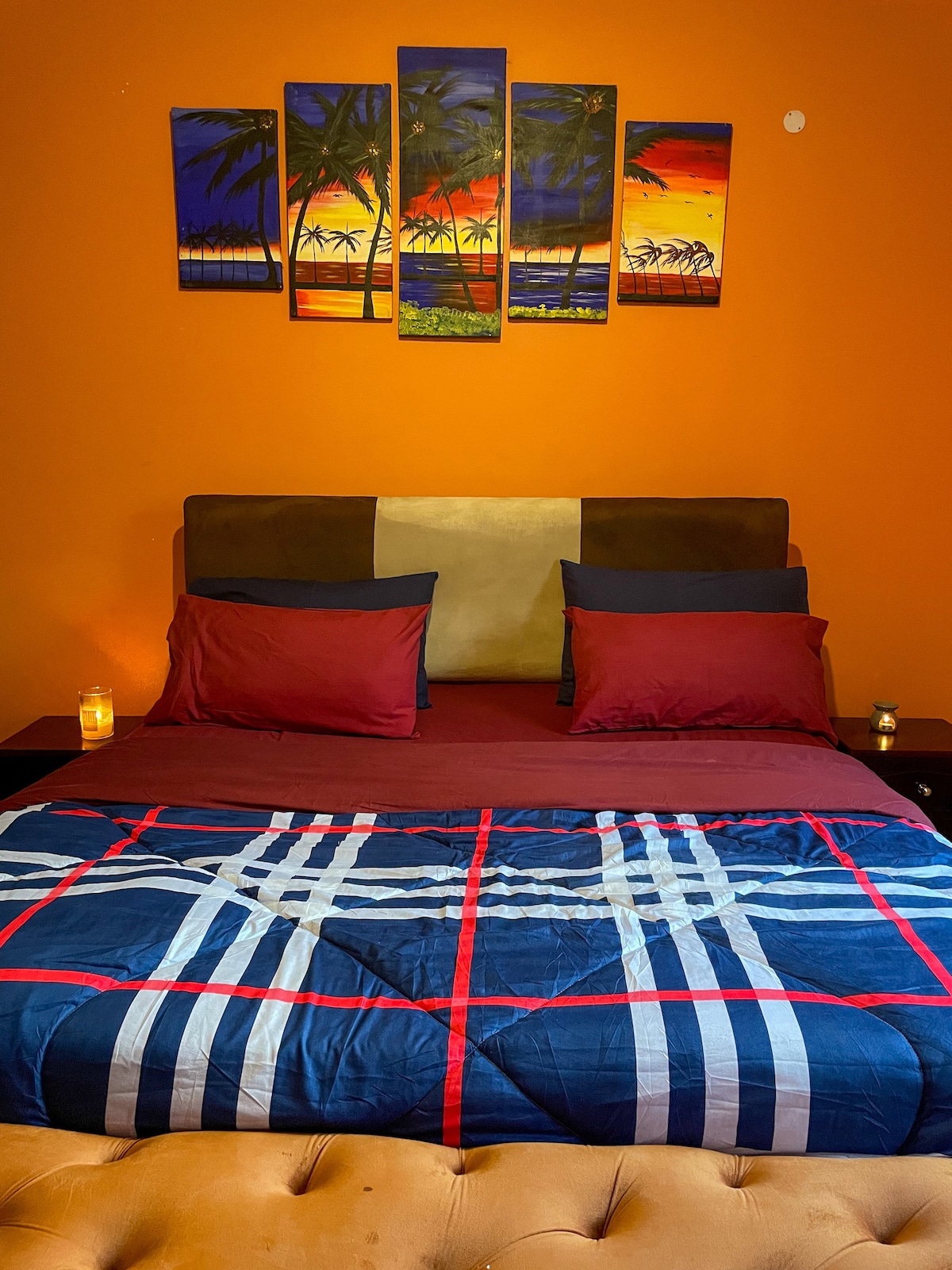 A master bedroom features a spacious bed adorned with a patterned quilt and decorative pillows. A vibrant wall art piece depicting a sunset over palm trees complements the warm orange wall. Soft lighting from nearby candles adds to the inviting atmosphere.