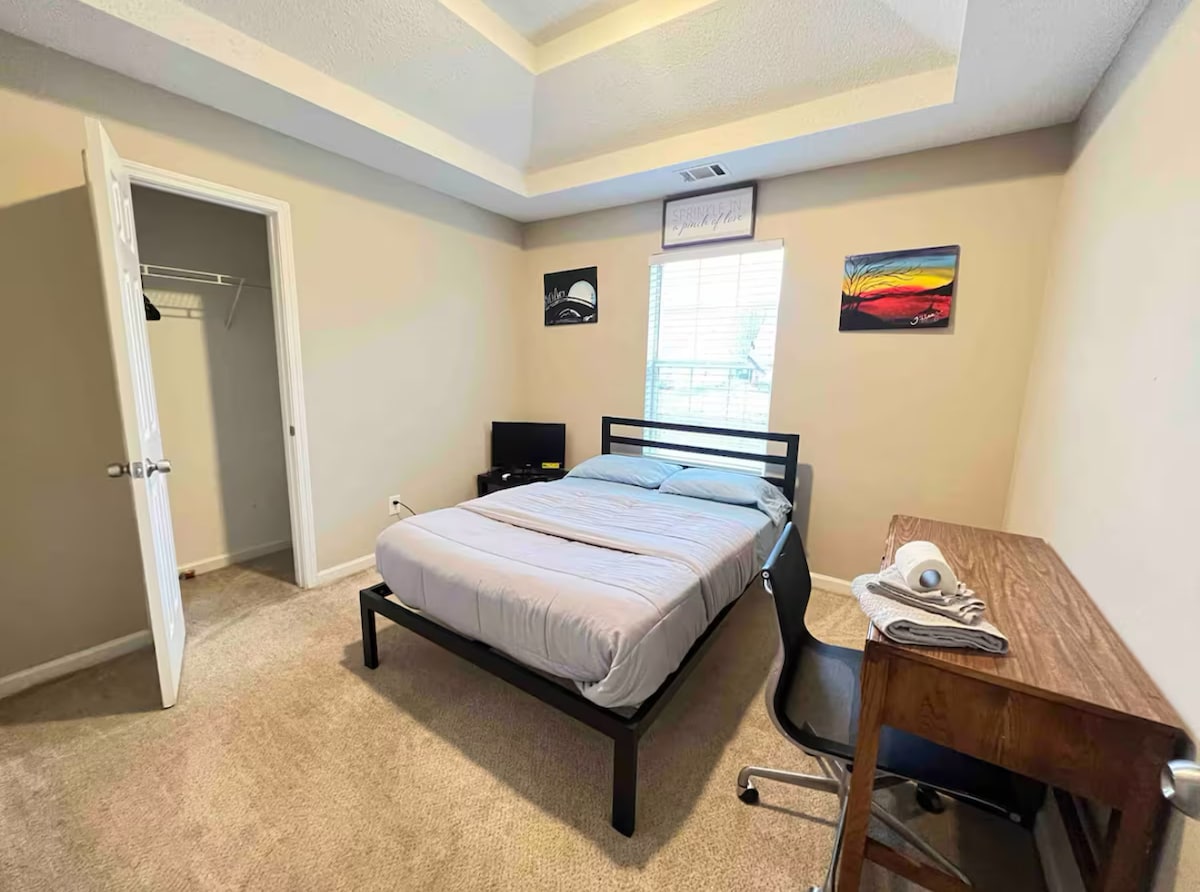 A simple bedroom features a spacious bed with light bedding. A small television is positioned on a stand. A desk and chair offer a workspace. A closet with sliding doors is visible, and natural light enters through the window, brightening the neutral-toned walls.