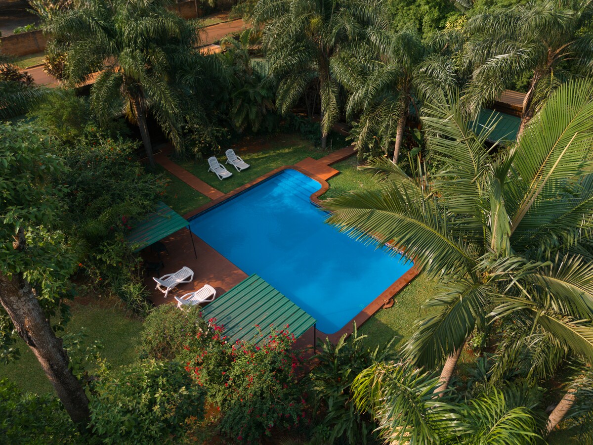 A spacious outdoor pool is surrounded by lush greenery and vibrant flowers. Sun loungers are positioned nearby on a wooden deck, offering a serene place to relax. The tranquil setting promotes a peaceful atmosphere in harmony with nature.