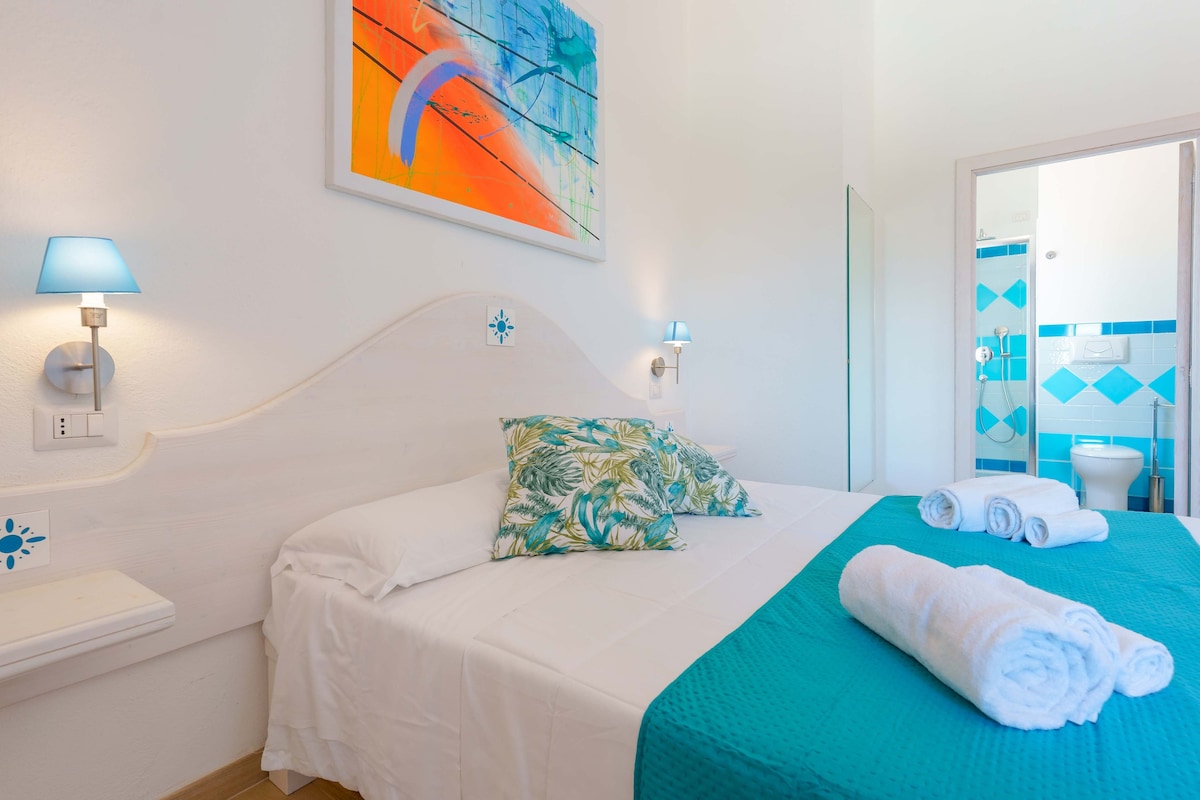 A bright room features a spacious bed with a light blue coverlet and decorative pillows. Walls are adorned with colorful artwork. Soft lighting is provided by wall-mounted lamps. An en-suite bathroom is visible in the background, showcasing blue and white tiled accents.