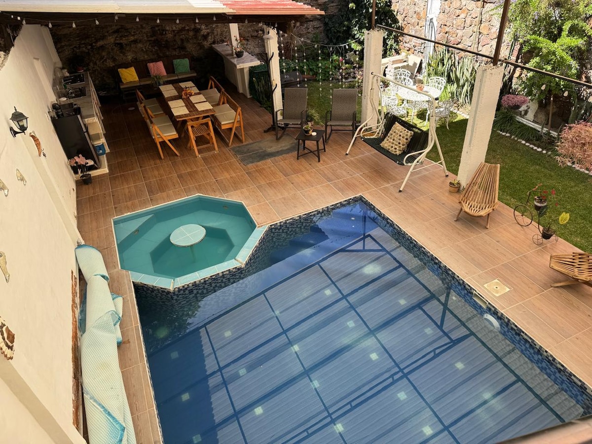 An outdoor area features a climate-controlled pool surrounded by tile flooring. Seating arrangements include a variety of chairs and tables under a shaded patio. Lush greenery and decorative elements create a relaxing atmosphere, with the pool's water reflecting natural light.