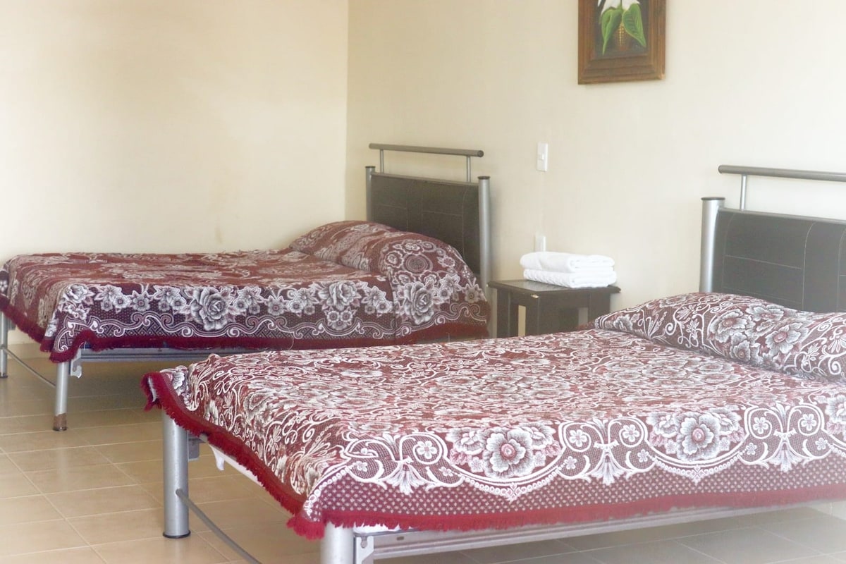 A spacious room is furnished with two large beds, each covered with a patterned red and white bedspread. A small bedside table is positioned between the beds, with neatly stacked towels visible on the side. An artistic piece hangs on the wall above.