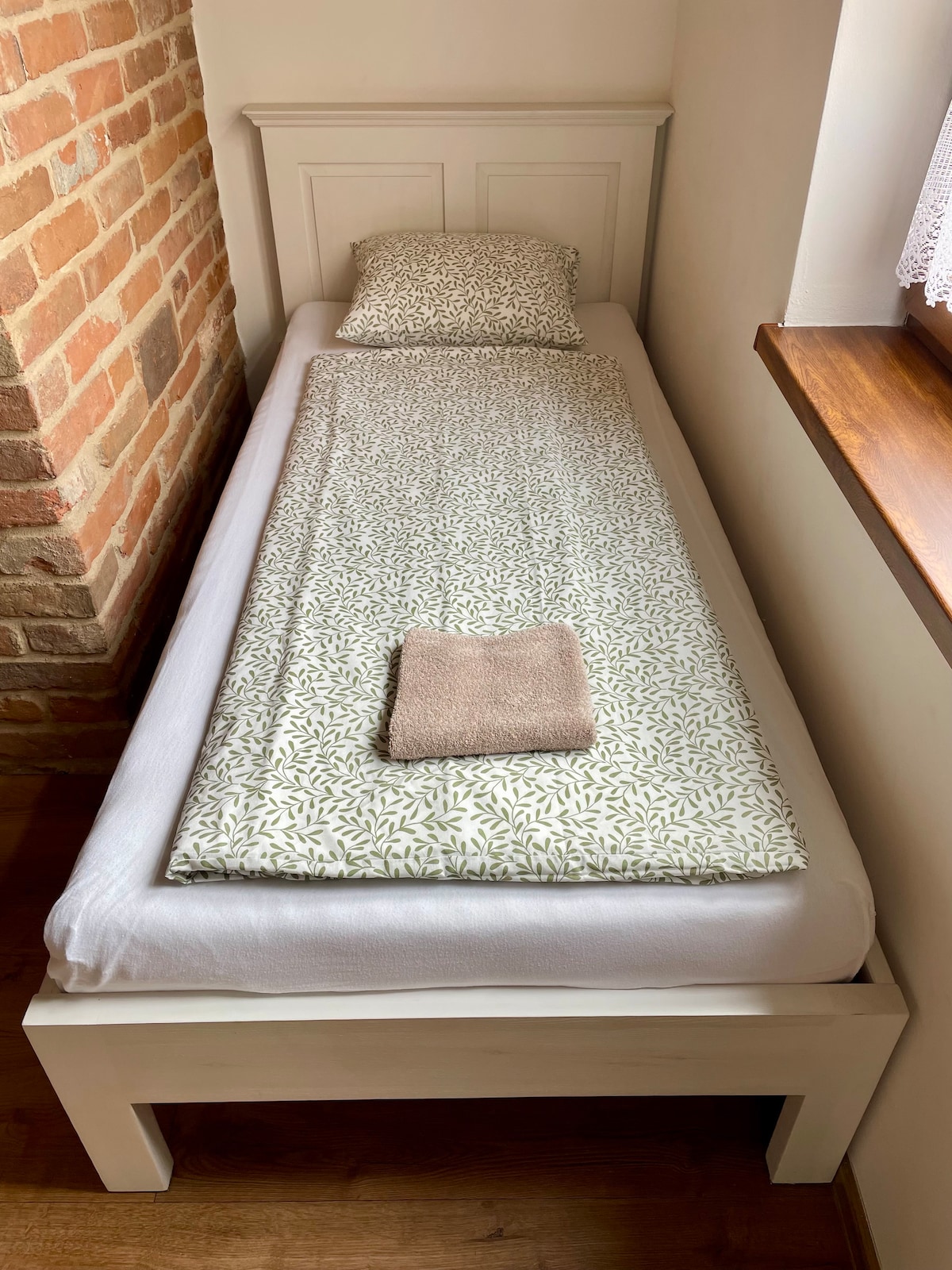 A single bed is positioned against a brick wall, featuring a floral-patterned bedspread and a soft pillow. A small towel is neatly placed on the bed, and natural light filters through a window, creating a subtle and calming environment.