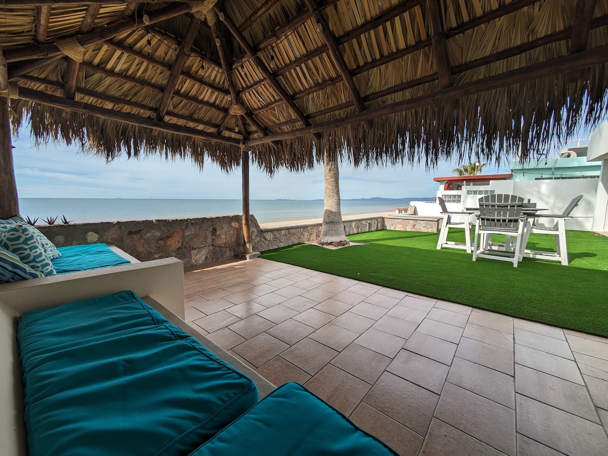 A shaded terrace beneath a thatched palapa features comfortable seating with turquoise cushions. A grassy area is visible, along with a white dining table and chairs. The serene beach and ocean can be seen in the background, creating a tranquil outdoor living space.