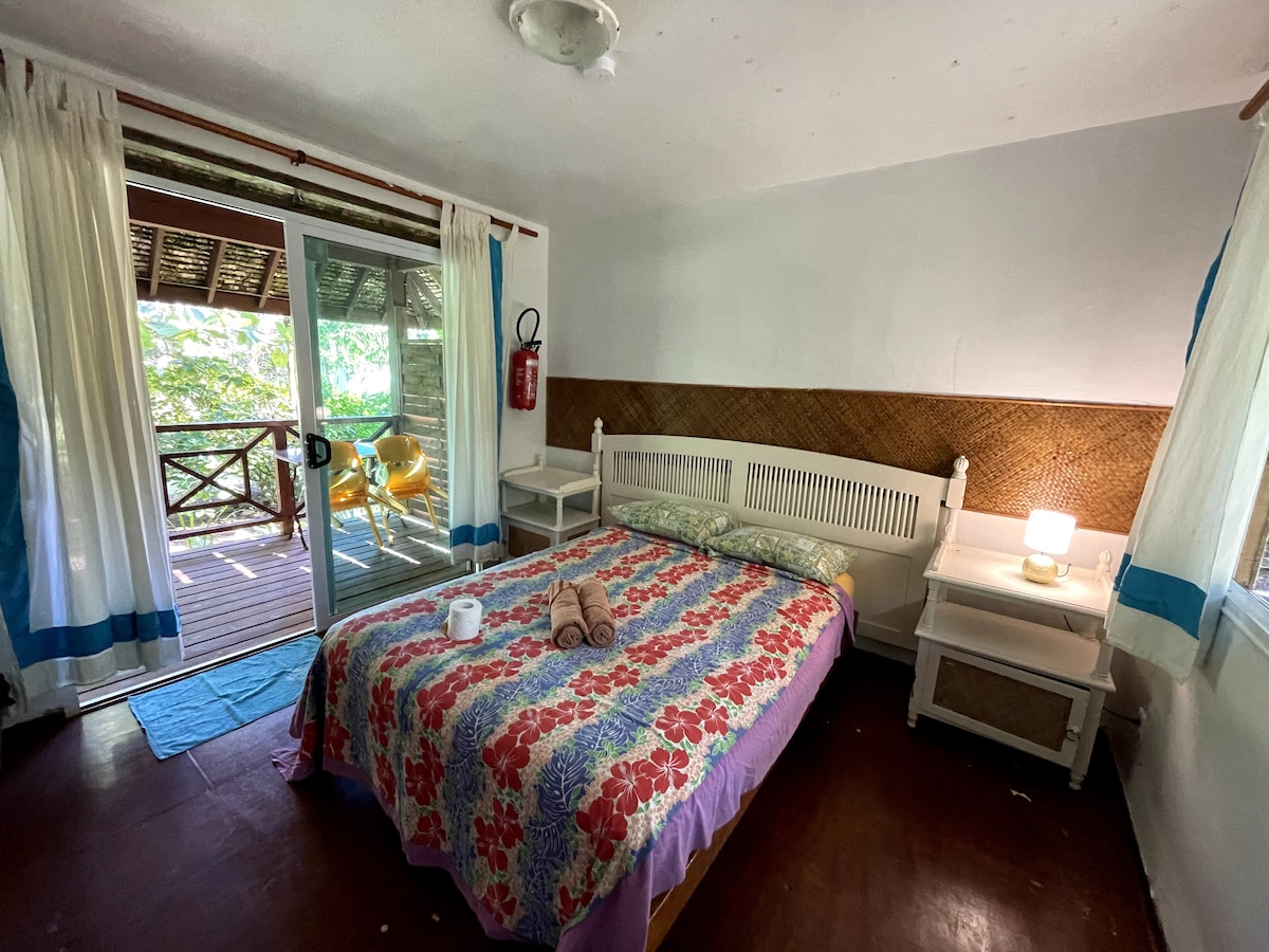 A bright sleeping area features a spacious bed adorned with a floral-patterned duvet. Two nightstands flank the bed, with a soft glow emanating from a bedside lamp. Light curtains frame the sliding glass doors that open to a private deck, revealing a serene outdoor space.