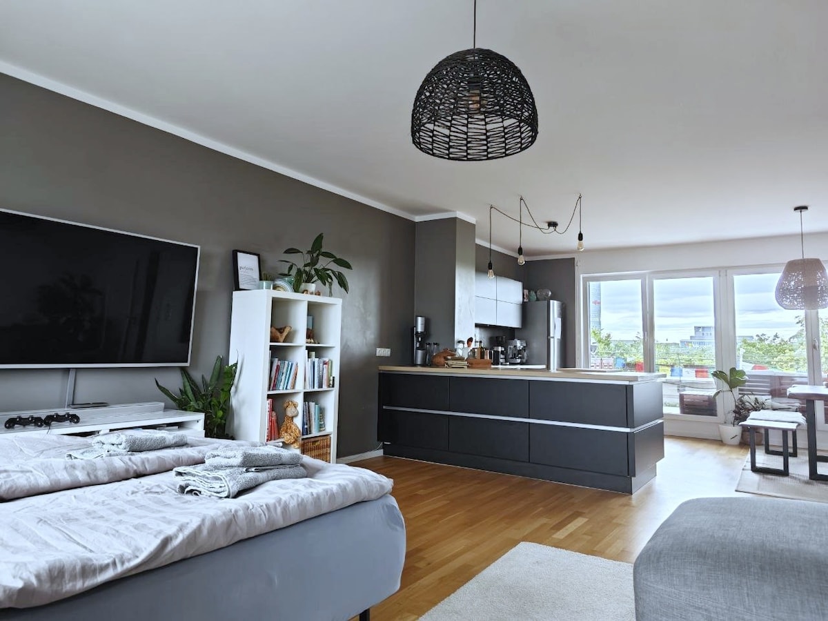 A spacious living area featuring a large flat-screen television mounted on a dark wall. An open kitchen with modern appliances is visible, complemented by a dining area bathed in natural light. Shelving units with plants and books add a touch of greenery and warmth.