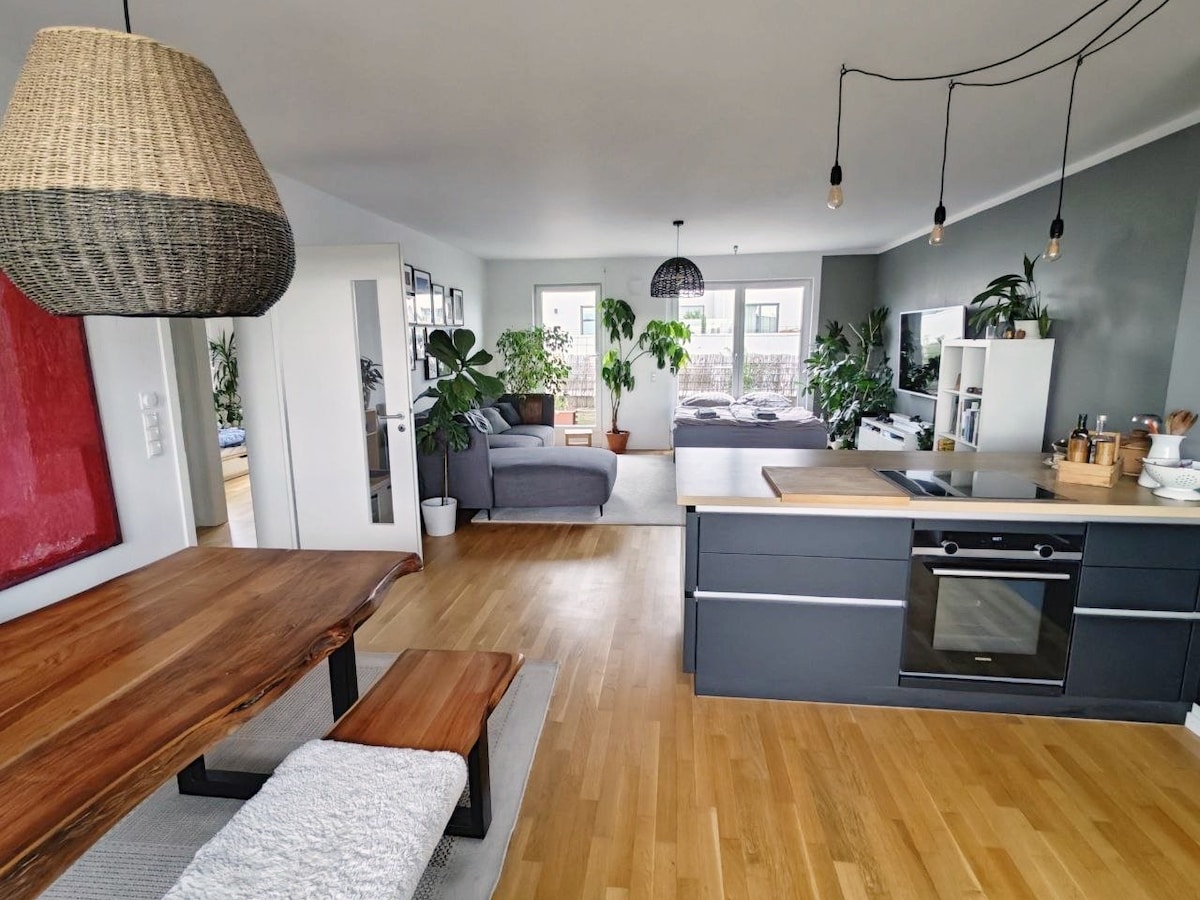 A modern kitchen area is observed with a wooden dining table and chairs, accompanied by soft seating in the adjacent living space. Greenery is featured throughout, and the room is illuminated by pendant lighting. Large windows provide a view into the outdoor terrace.