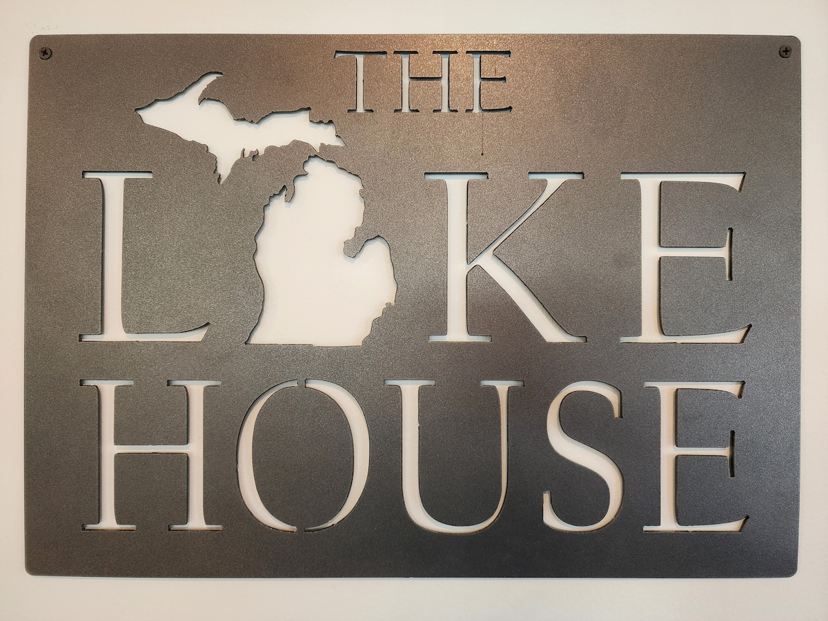A metal sign is displayed on a wall, featuring the words 'THE LAKE HOUSE' alongside an outline of the state of Michigan. The design highlights a local connection to the lakeside environment.