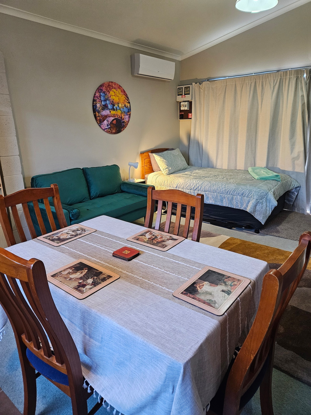 A cozy living space is furnished with a green sofa and a spacious bed draped in a light blue cover. A dining table with four wooden chairs is set with placemats. A colorful wall art piece adds visual interest, while curtains softly separate areas.