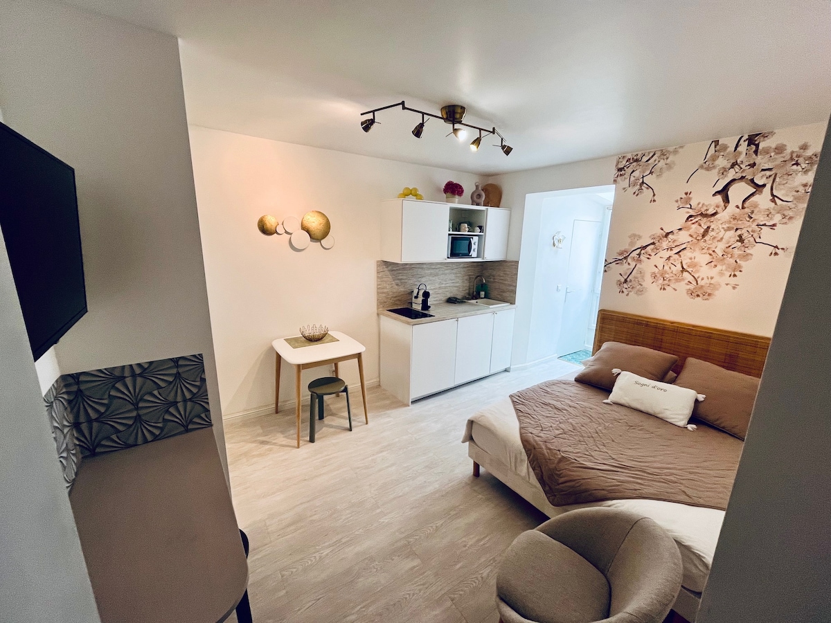 The studio is equipped with a spacious bed, a kitchenette, and a small dining area. A wall-mounted television is visible, and natural light brightens the interior through the door leading to a private bathroom. Decorative elements include wall art and minimalist furnishings.