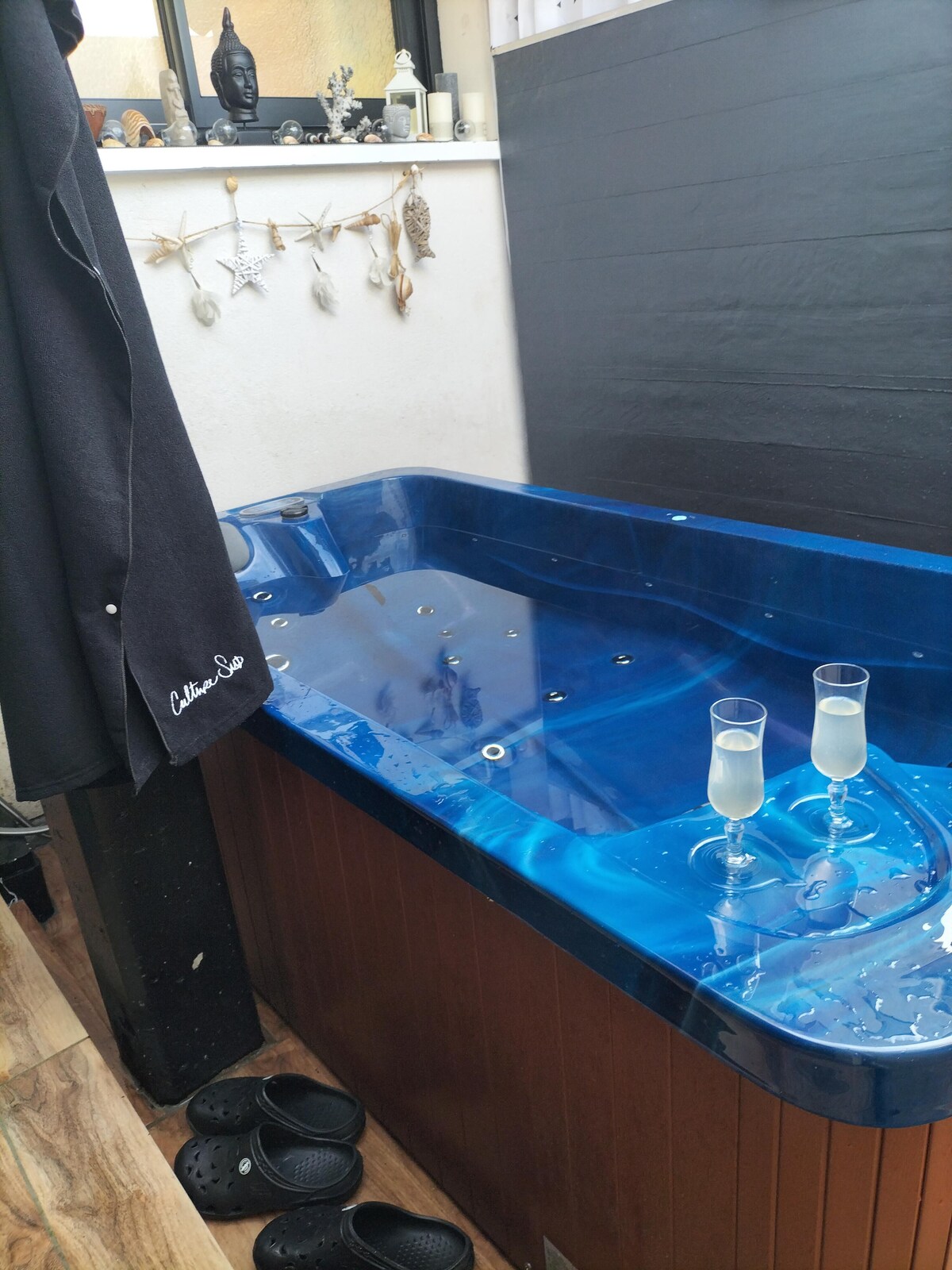 A blue spa tub features a series of jets and is positioned against a wooden backdrop. Two glasses are placed on the edge of the tub, resting on a flat surface. Black footwear is seen nearby, enhancing the relaxing ambiance.