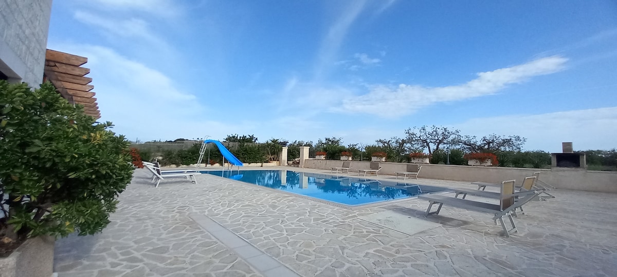 An outdoor area features a large infinity pool surrounded by stone decking. Loungers are positioned around the pool for relaxation. A slide is visible for added fun. Lush greenery and flowering plants enhance the serene landscape, with a gravel path leading to different seating areas.