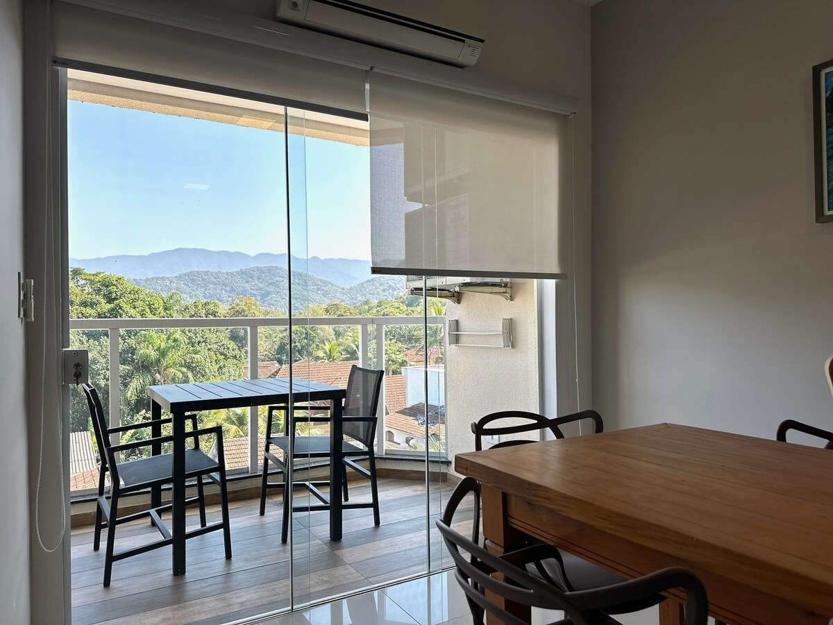 A sliding glass door opens to a balcony with a scenic view of lush mountains and greenery. The interior features a dining area with a wooden table and black chairs, complemented by natural light from the large window.