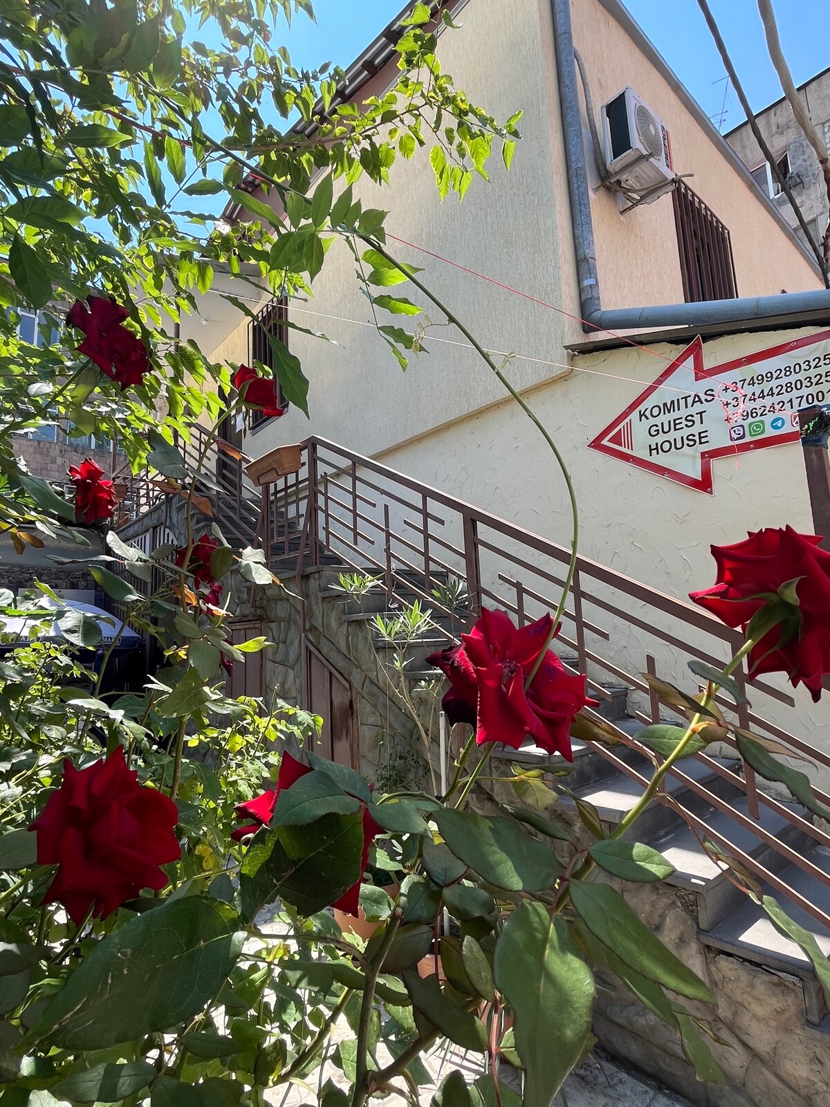 The entrance to the guest house is framed by vibrant red roses, with a stairway leading up to the building. A sign indicating the guest house is visible, and air conditioning units are mounted on the exterior wall.