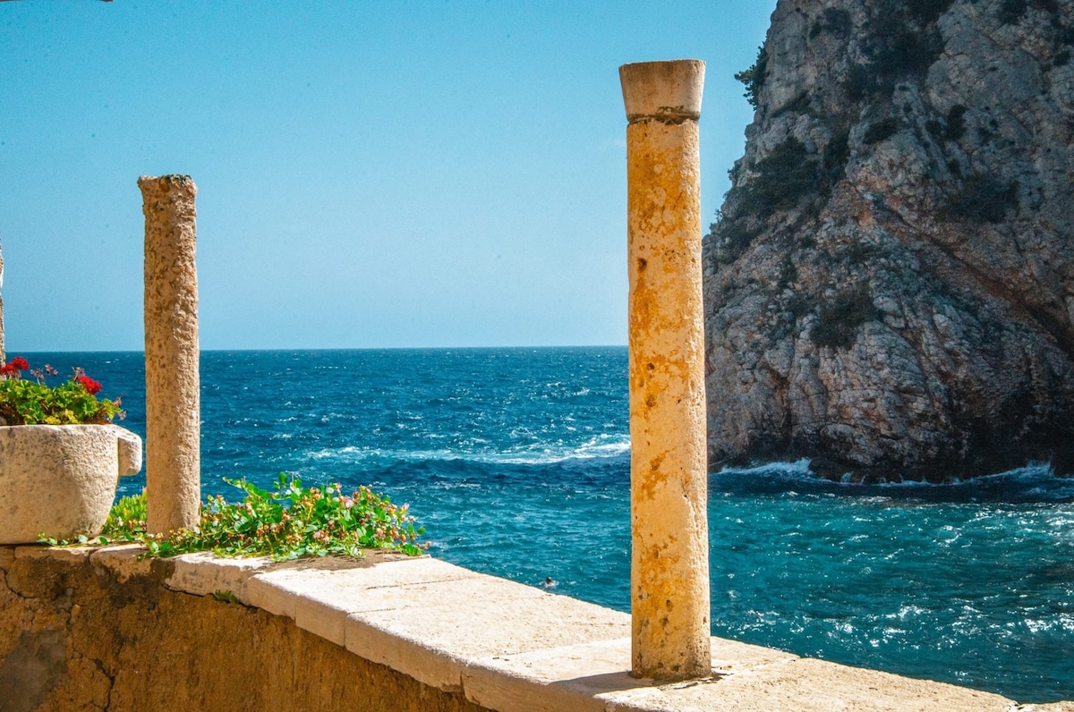 A view captures the vibrant blue waters of the Adriatic Sea, framed by two weathered stone columns. The rocky cliffside rises in the background, adorned with greenery, while the horizon meets the clear sky, creating a serene coastal atmosphere.