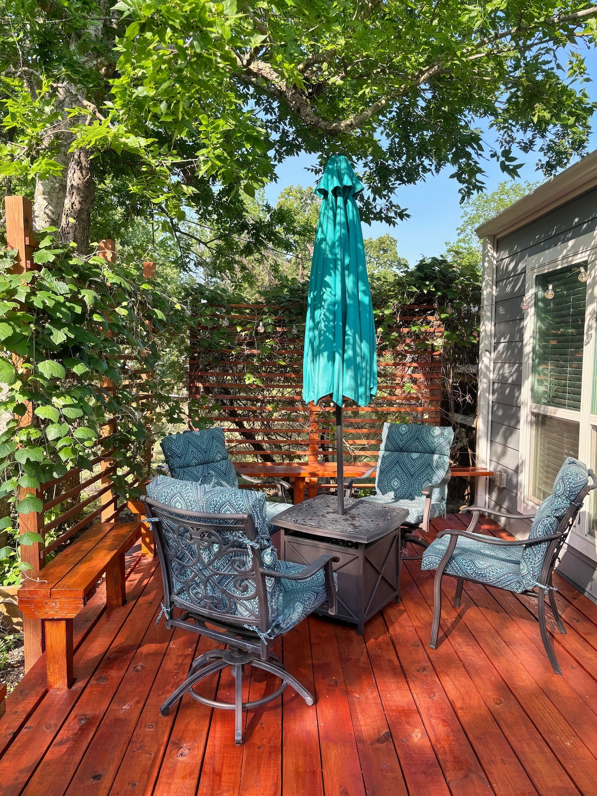 A shaded deck area features a round table with a central umbrella, surrounded by four cushioned chairs. A wooden bench is positioned against a backdrop of green foliage and wood lattice, creating a relaxing outdoor space.